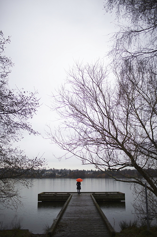 A woman standing at the end of a dock with an orange umbrella on a cloudy, grey day in Seattle, Seattle, Washington, USA