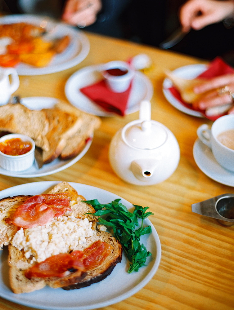 A table covered with plates of food. A cooked breakfast of bacon, eggs and toast and pot of tea. Two people seated , England