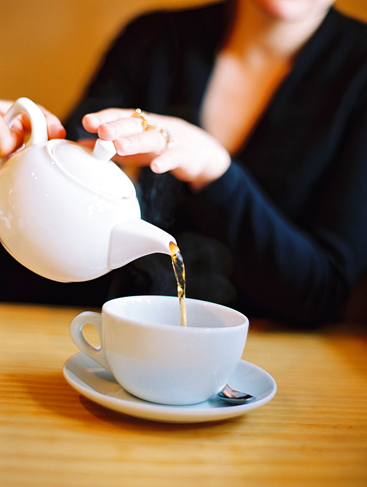 A woman seated pouring a cup of tea out of a white china tea pot, England