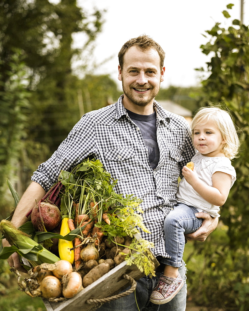 Man standing in his allotment with his daughter, smiling, holding a box full of freshly picked vegetablesAllotment, England