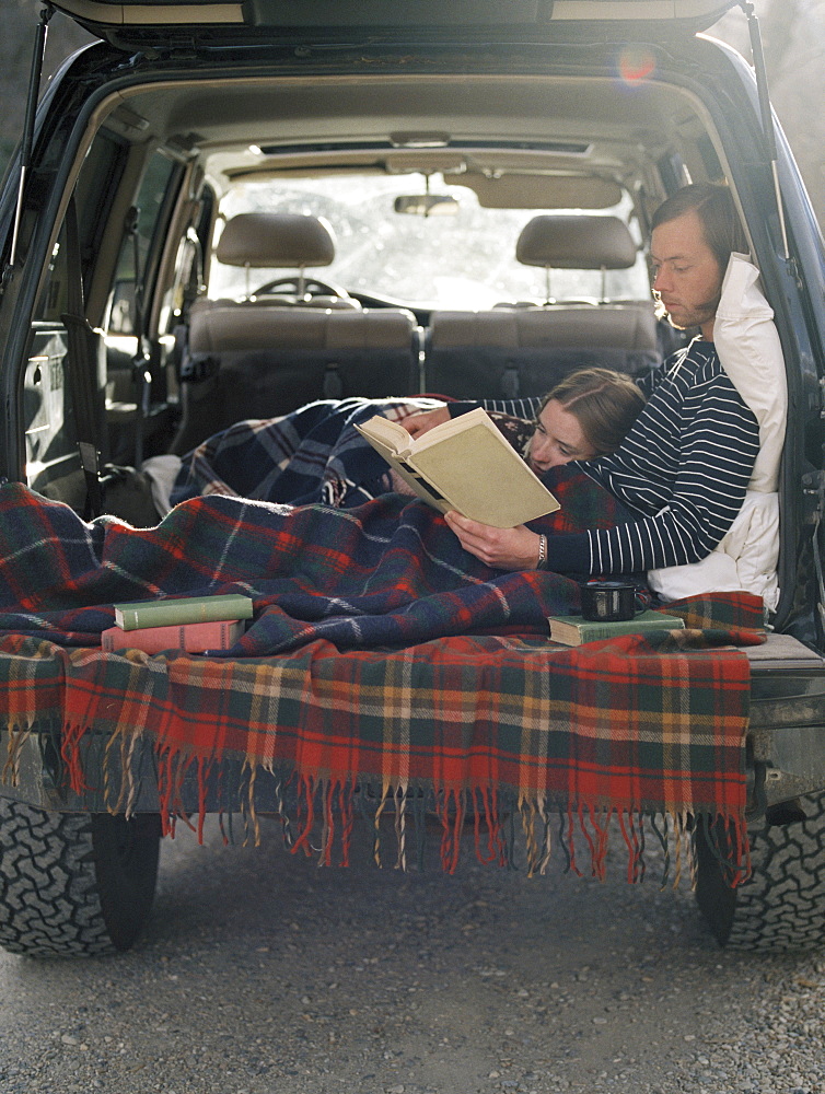 Young couple lying in the back of their car, reading a book, Millcreek, Utah, United States of America