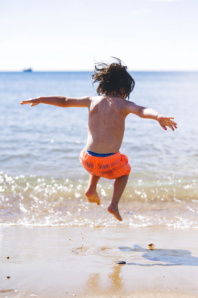 A boy in swimming trunks jumping over waves on the sea shore, England