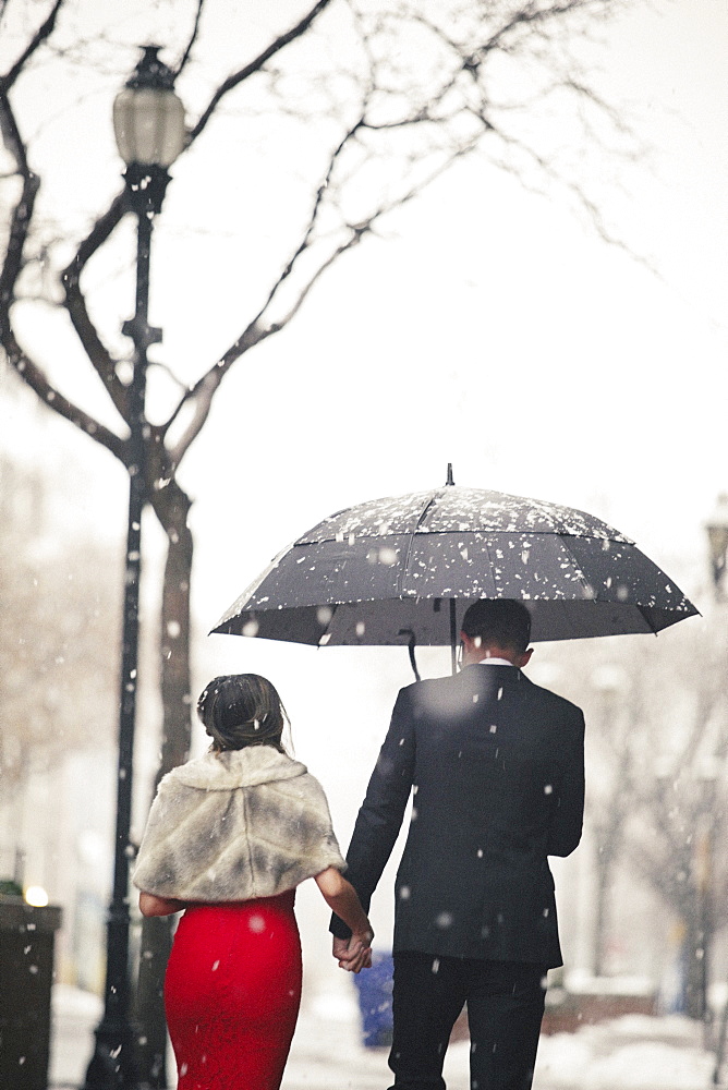 A woman in a long red evening dress and a man in a suit, walking through snow in the city.