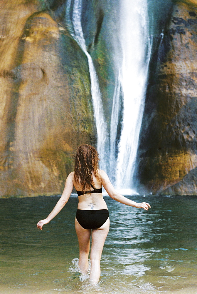 Woman in a black bikini wading into a pool with a waterfall cascade tumbling down a cliff, United States of America