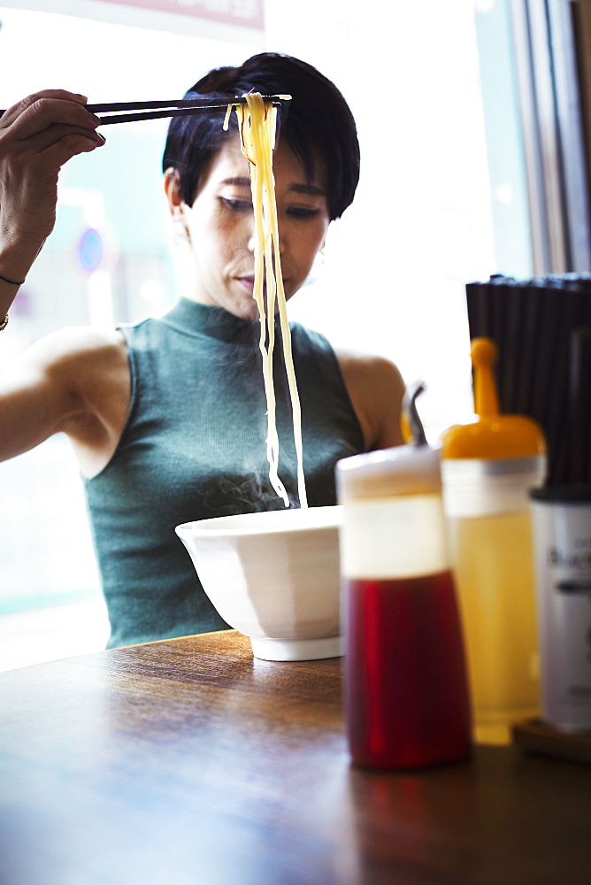 A ramen noodle cafe in a city. A woman seated eating a ramen noodle dish using chopsticks, Japan