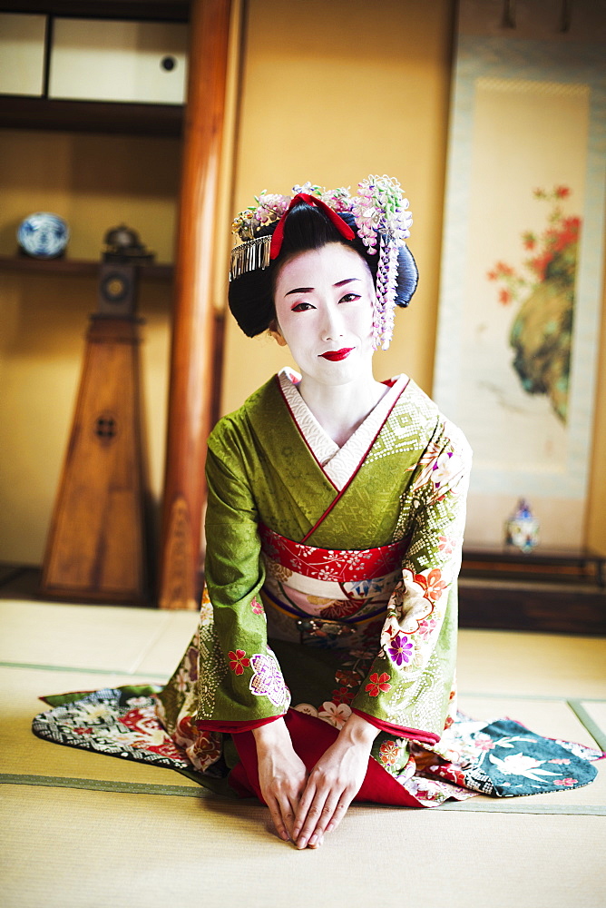 A woman dressed in the traditional geisha style, wearing a kimono and obi, with an elaborate hairstyle and floral hair clips, with white face makeup with bright red lips and dark eyes. Kneeling on the floor, Japan