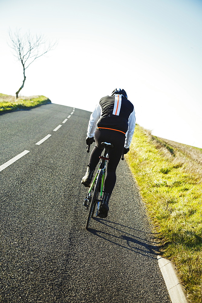 A cyclist pedalling along a country road, rear view.