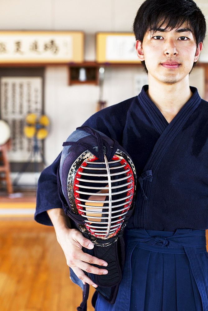 Male Japanese Kendo fighter standing in a gym, holding Kendo mask, looking at camera, Kyushu, Japan