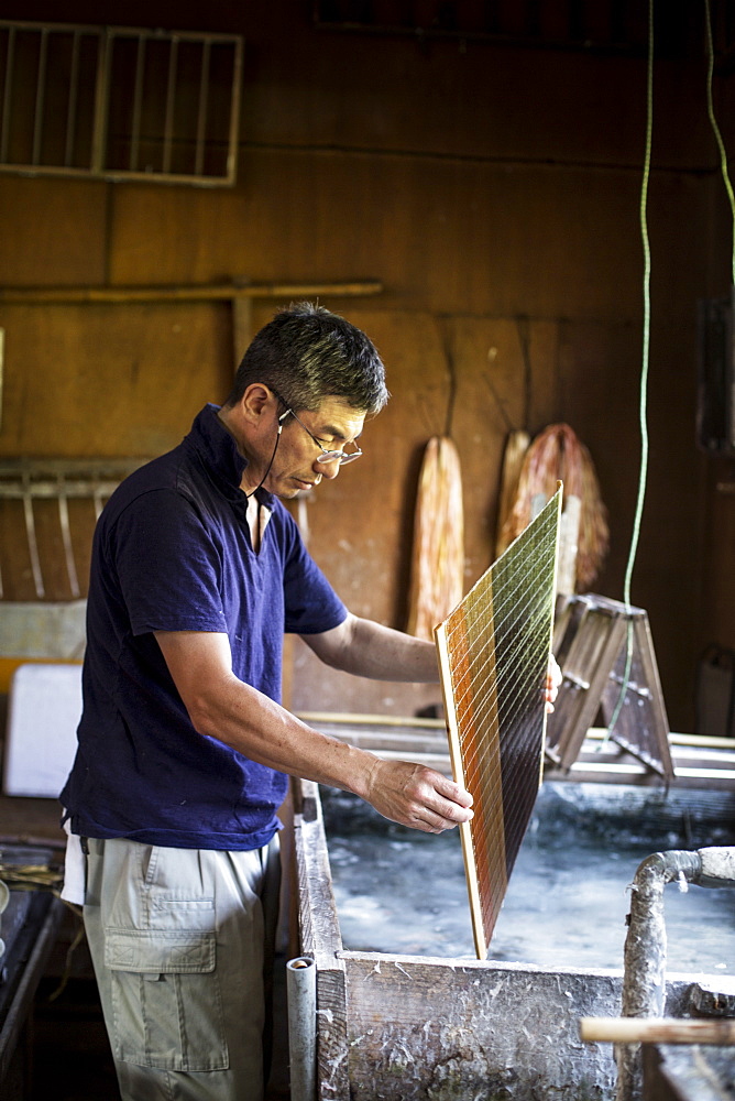 Japanese man in a workshop holding a wooden frame of dried pulp over a basin of water, making traditional Washi paper, Kyushu, Japan