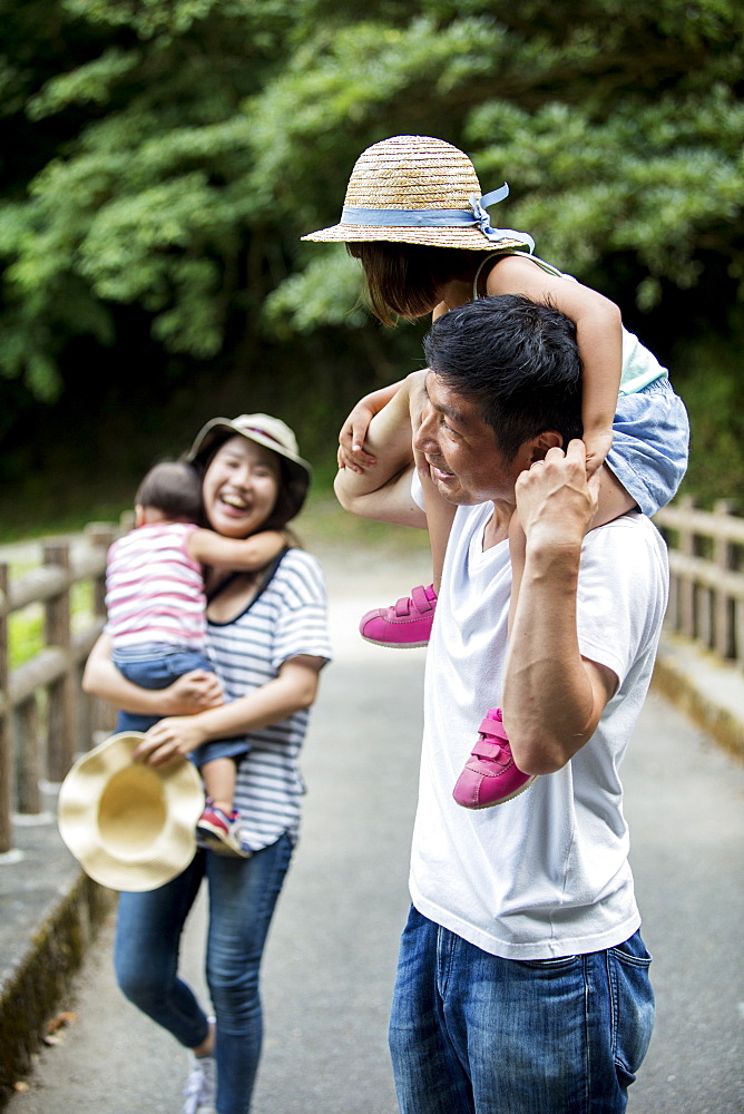 Japanese girl, smiling woman holding hat and man carrying toddler on his shoulders standing on wooden bridge, Kyushu, Japan