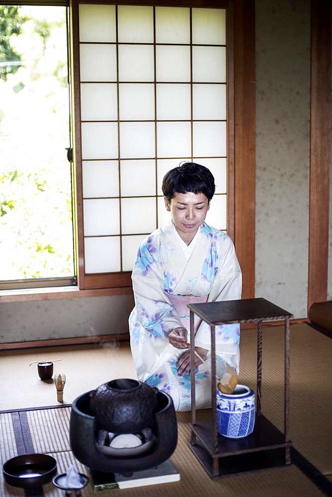 Japanese woman wearing traditional white kimono with blue floral pattern kneeling on tatami mat during tea ceremony, Kyushu, Japan