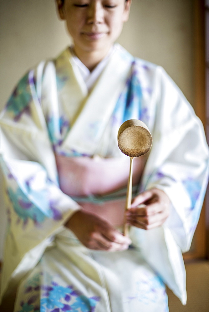 Japanese woman wearing traditional white kimono with blue floral pattern kneeling on floor during tea ceremony, holding a Hishaku, a bamboo ladle, Kyushu, Japan