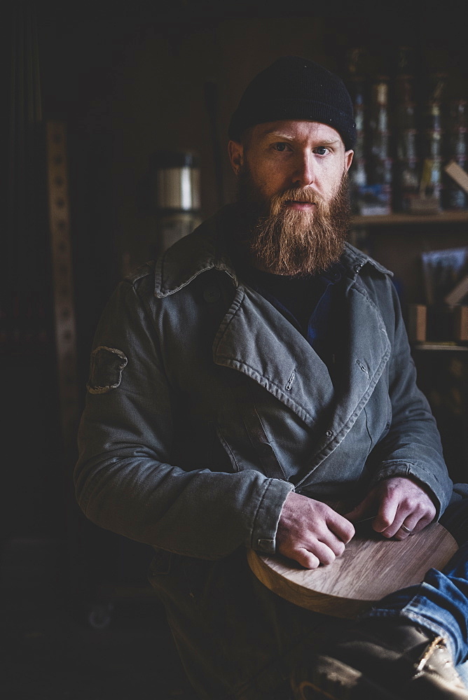 Bearded man wearing black beanie sitting in workshop, holding piece of wood, looking at camera, Berkshire, England