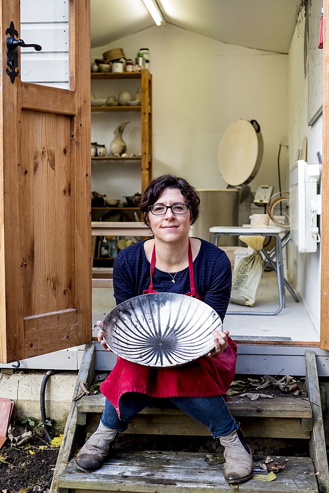 Woman wearing red apron sitting on steps outside her workshop, holding ceramic bowl with black line pattern, England