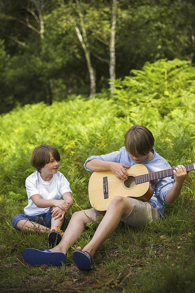 Two boys camping in New Forest. Sitting on the grass, one playing a guitar, Hampshire, England