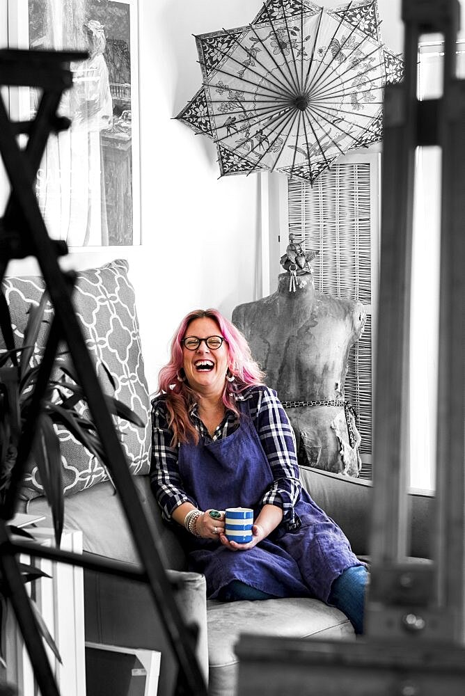 Laughing woman with long blond wavy hair with pink streaks sitting on a sofa, looking at camera, Oxfordshire, England