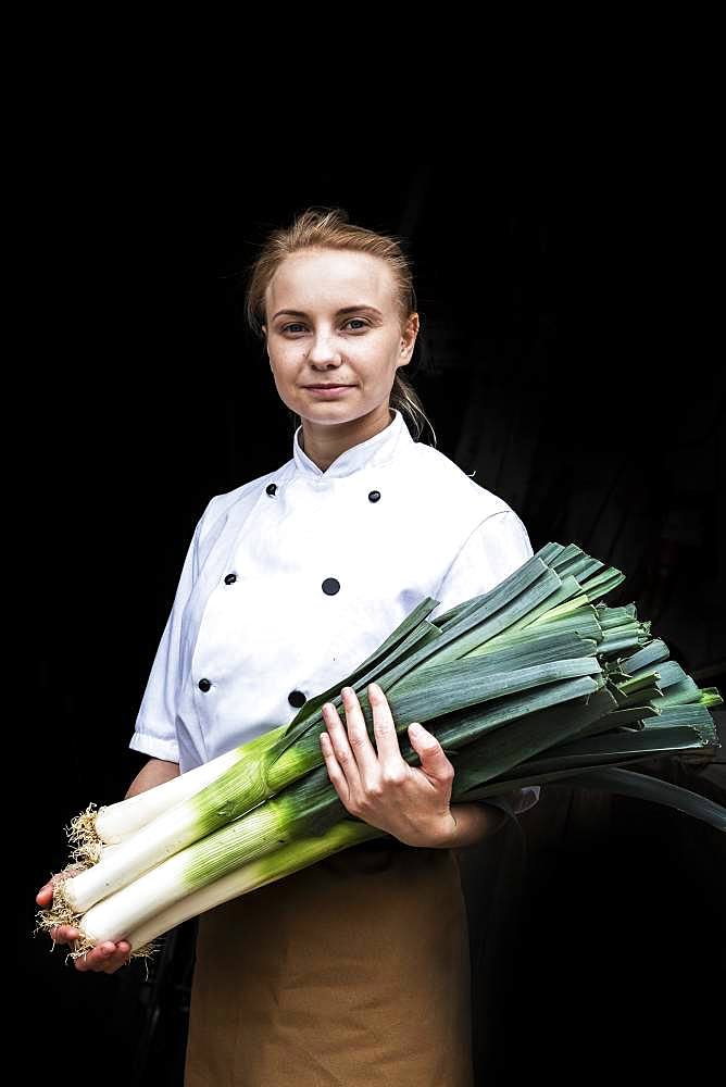 Woman wearing chef's jacket standing indoors, holding bunch of leeks, looking at camera