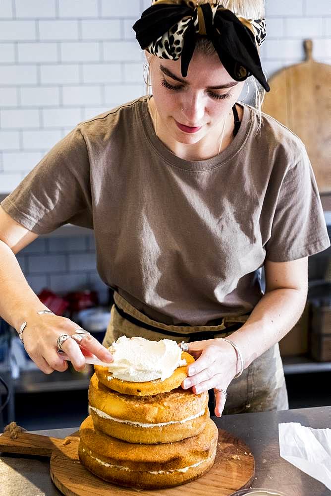 A cook working in a commercial kitchen assembling a layered sponge cake with fresh cream