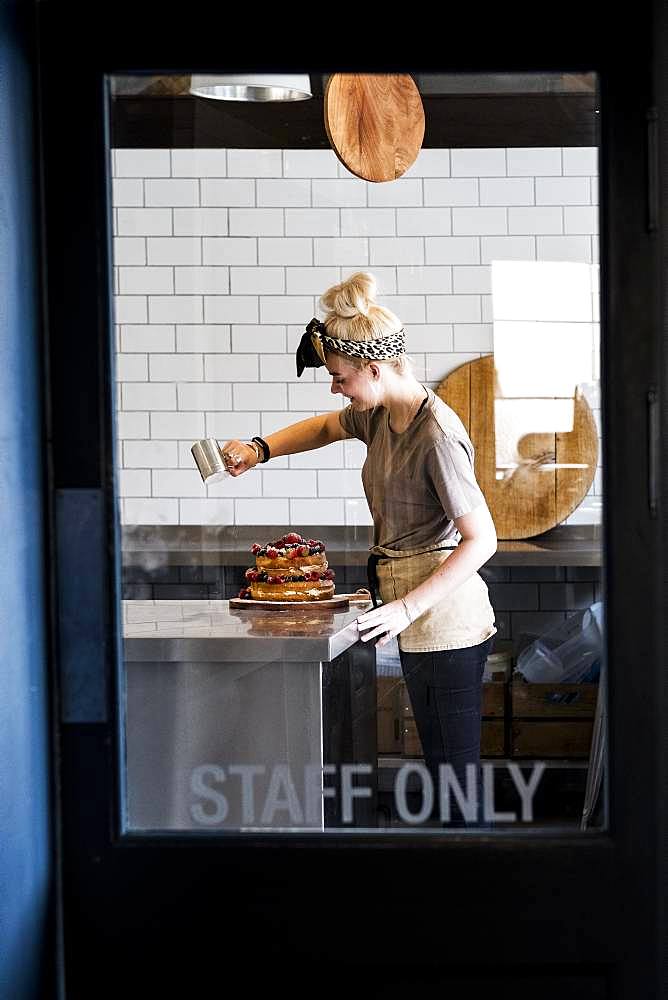 View through a door marked Staff Only, of a cook working in a commercial kitchen sprinkling icing sugar over a layered cake with fresh fruit