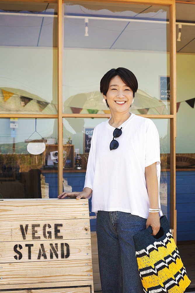 Japanese woman standing outside a farm shop, holding shopping bag, smiling at camera, Kyushu, Japan