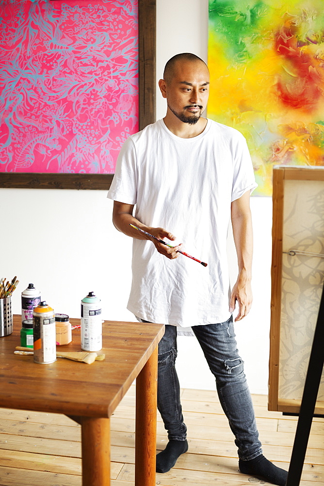 Japanese man standing in art gallery, holding paintbrush, looking at artwork on easel, Kyushu, Japan