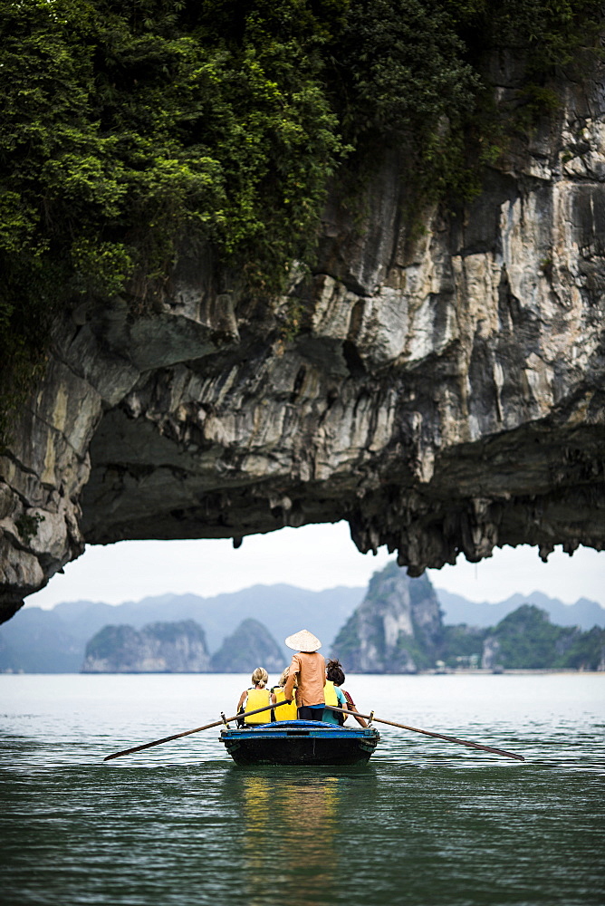 Rear view of man wearing straw hat transporting small group of people on a boat, rowing underneath natural rock arch, Vietnam