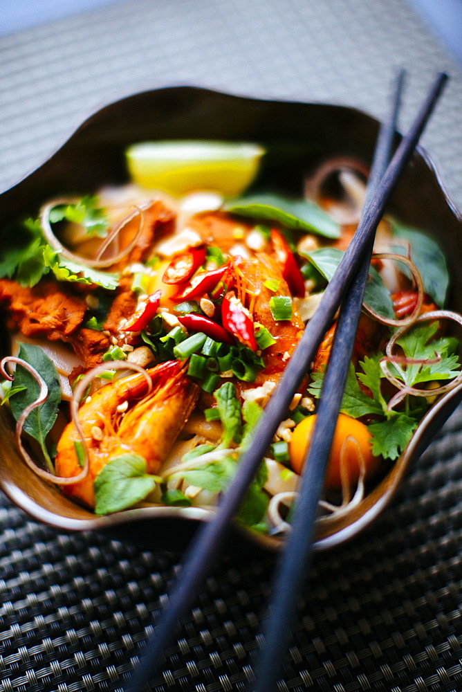 High angle close up of chopsticks on bowl with Asian food containing noodles, prawns, vegetables and chili garnish, Vietnam