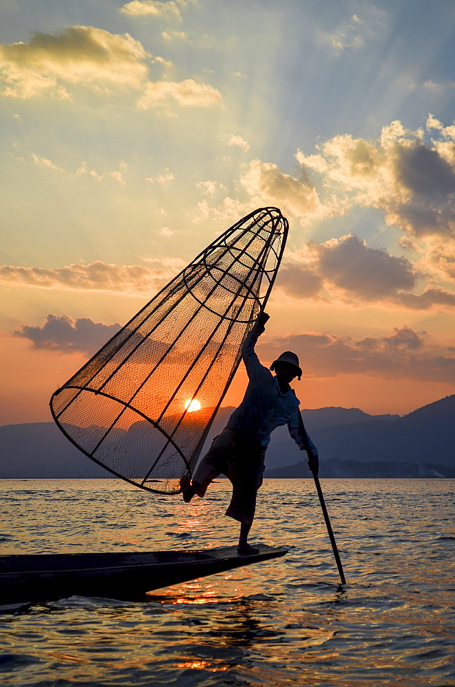 Traditional fisherman balancing on one leg on a boat, holding fishing basket, fishing on Lake Inle at sunset, Myanmar