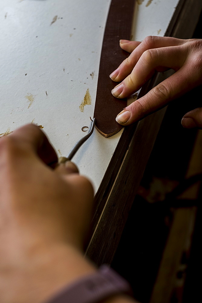 High angle close up of person working in saddler's workshop, using hand tool on leather strap, Berkshire, United Kingdom