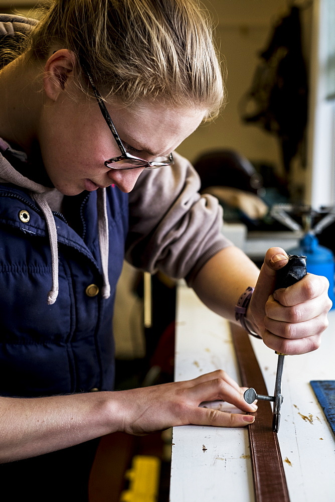 Female saddler standing in workshop, using hand tool on leather strap, Berkshire, United Kingdom