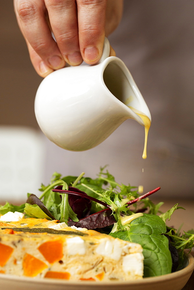 Close up of person drizzling dressing onto baby leaf salad and frittata in a cafe