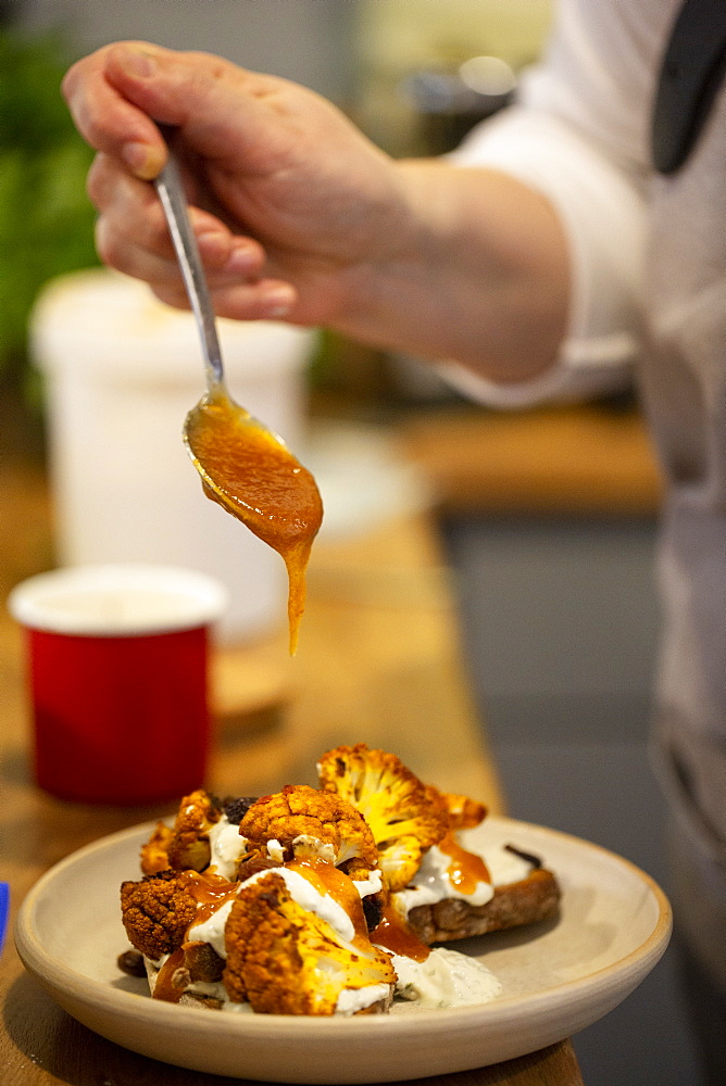 Close up of person drizzling mango chutney onto roasted cauliflower with yoghurt and sesame seeds on toasted sourdough bread in a cafe