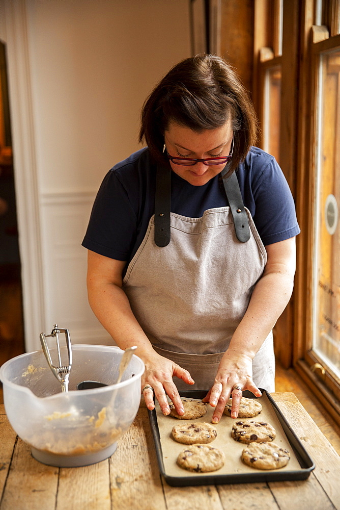 Woman wearing glasses and apron standing at wooden table, baking chocolate chip cookies