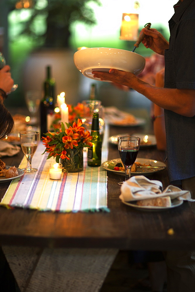 Person standing at a table holding a bowl, wine glasses, plates, flowers and candles on the table