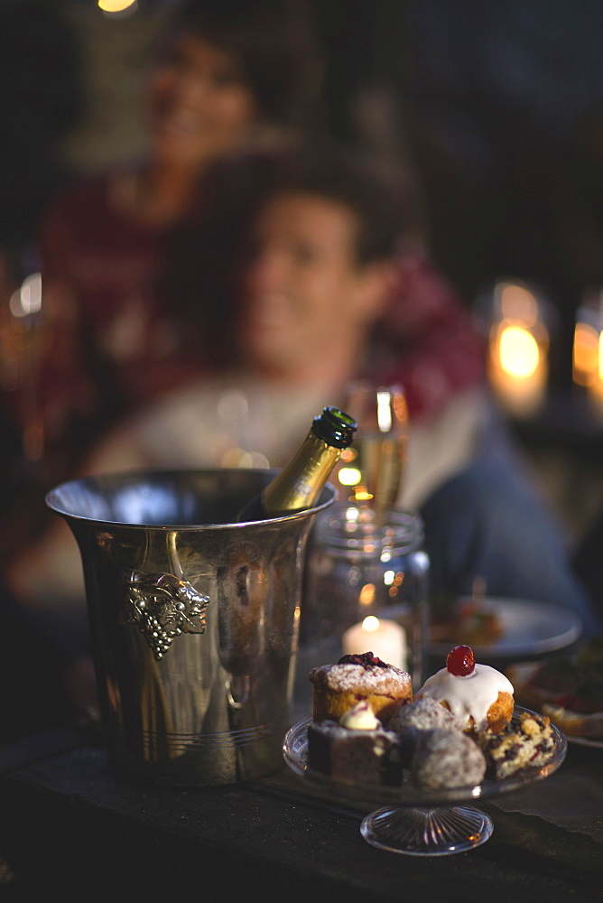 Close up of champagne bottle in metal wine cooler, glass cake stand with selection of cakes, person in the background