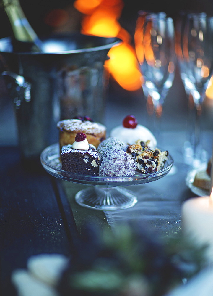 Close up of glass cake stand with a selection of cakes, champagne glasses and wine cooler in the background