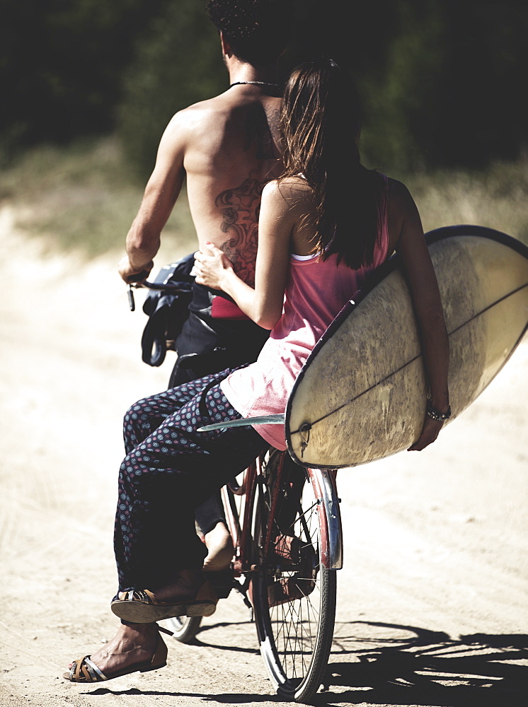 Rear view of man and woman on a bicycle, woman sitting on rack, holding a surfboard