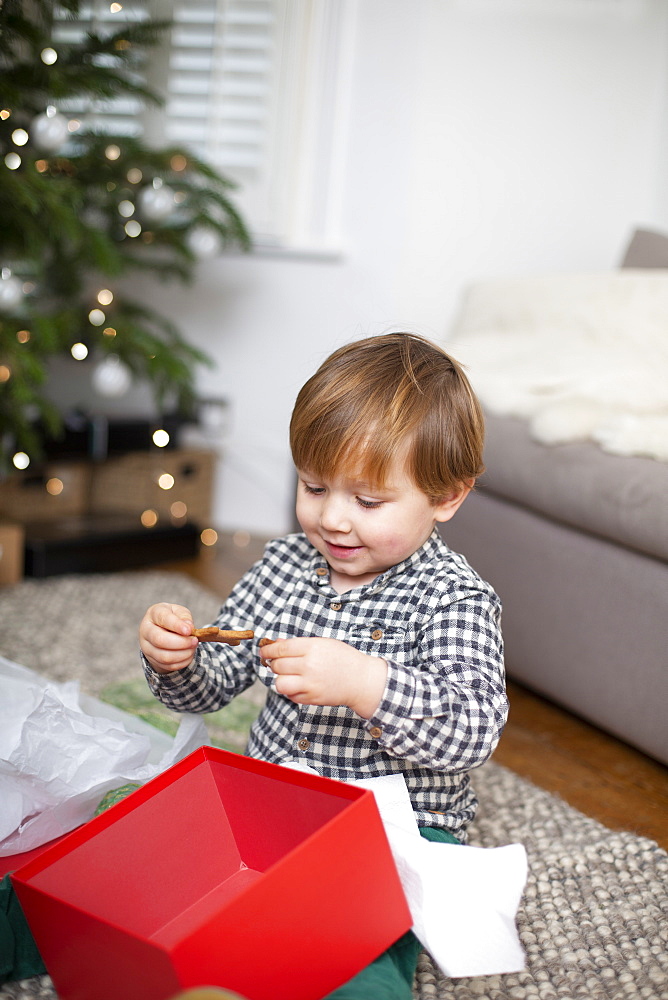 Young boy sitting on living room floor, unwrapping Christmas present in red box