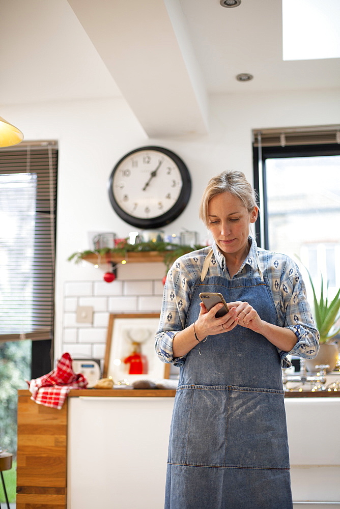 Blond woman wearing blue apron standing in kitchen, using mobile phone