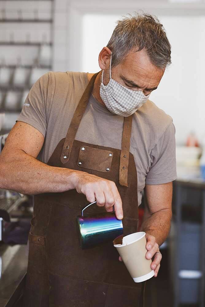 Male barista wearing brown apron and face mask working in a cafe, pouring coffee