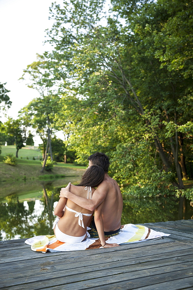 A young couple, boy and girl, sitting close together on a wooden jetty by a water pool, Maryland, USA