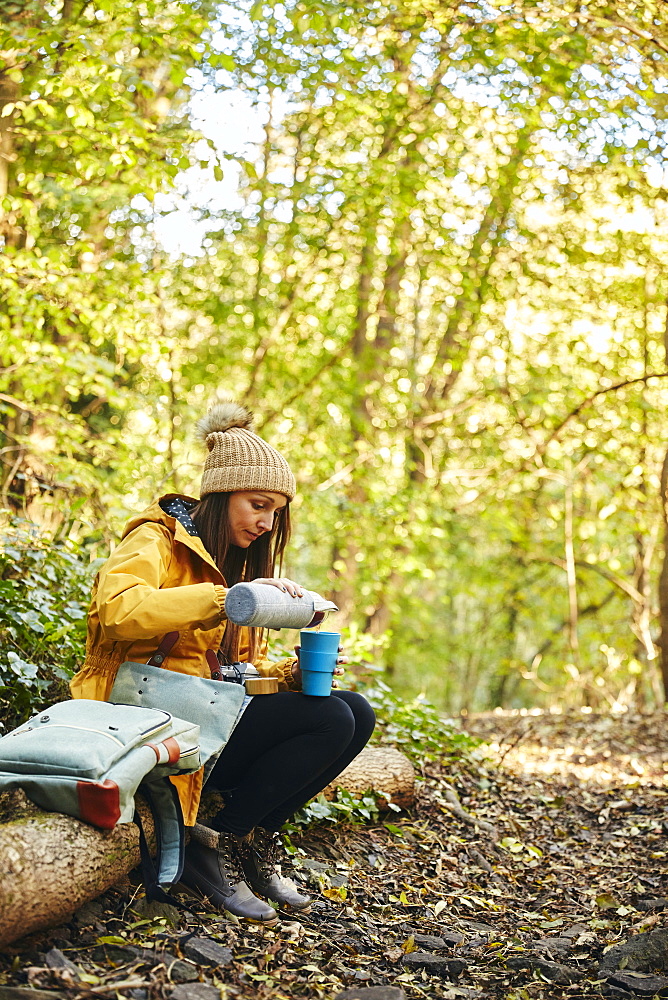 Woman sitting on tree trunk in woodland pouring drink from thermos flask