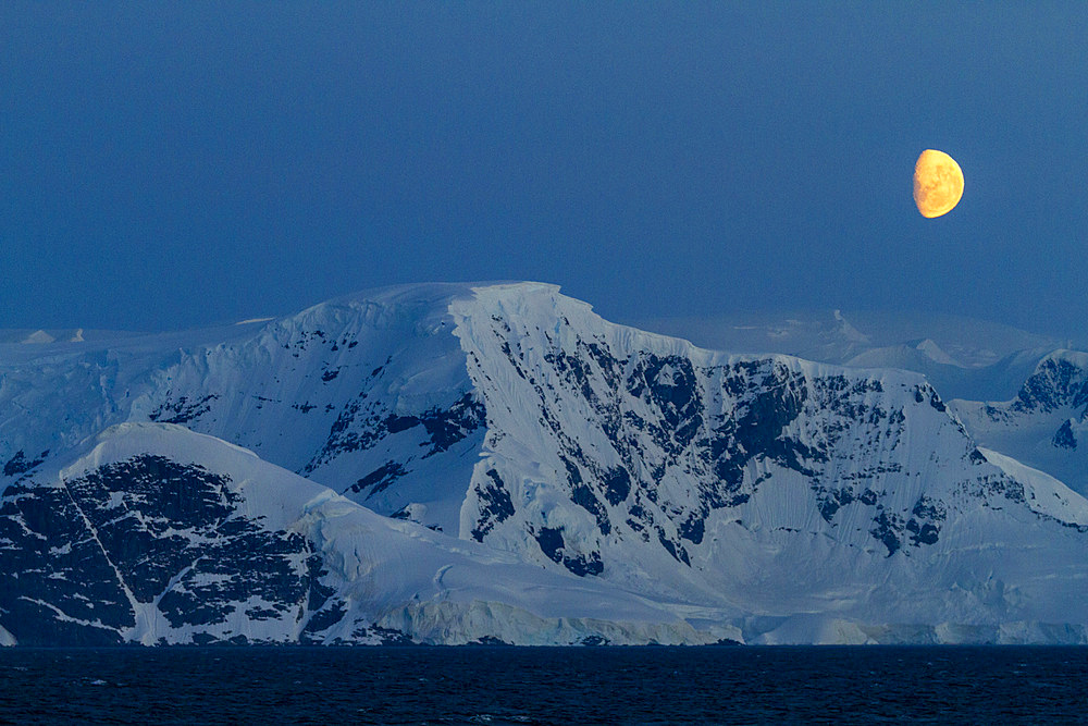 View of the nearly full moon rising over snow-covered mountains on the Antarctic Peninsula, Antarctica.