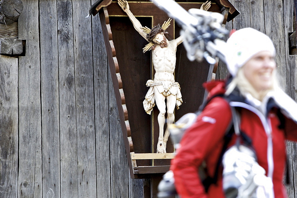 Young female skier walking past a wayside cross, See, Tyrol, Austria