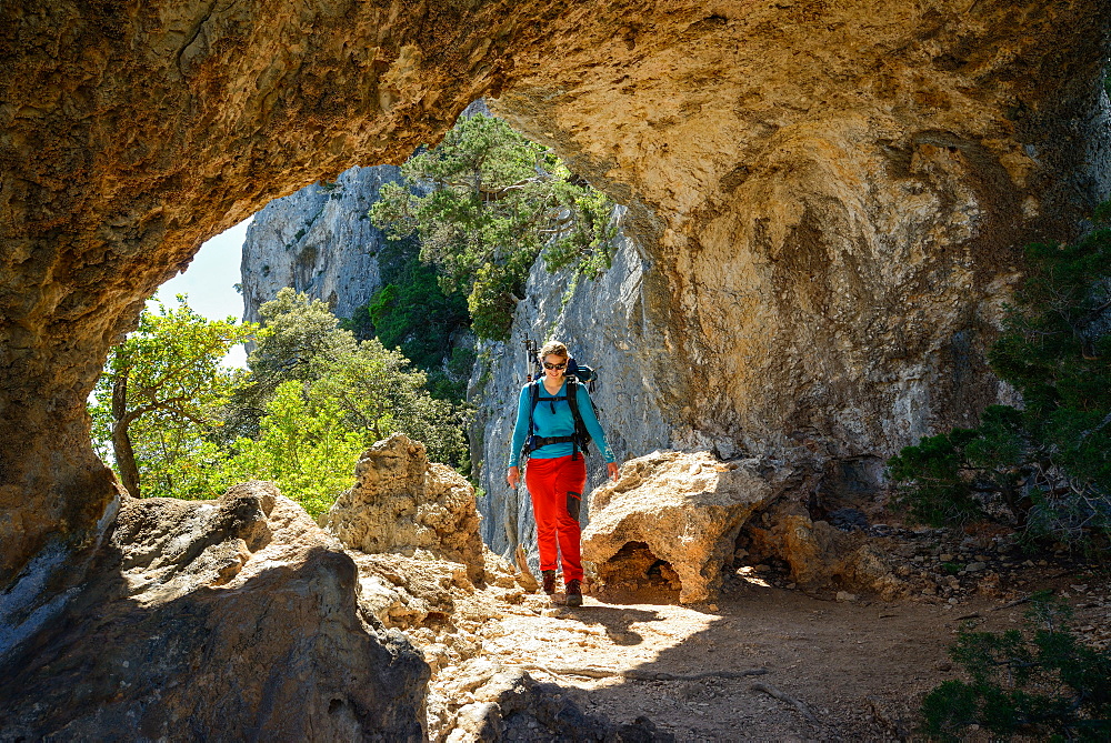 A young woman with trekking gear hiking through the rock arch Arcu su Feilau at the mountainous coast above the sea, Golfo di Orosei, Selvaggio Blu, Sardinia, Italy, Europe