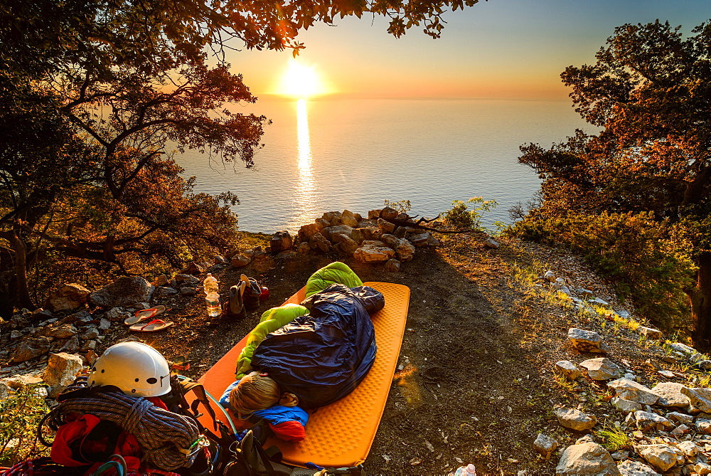 Young woman sleeping in her sleeping bag at sunrise near the bay Cala Biriola, Trekking- and climbing gear visible, Golfo di Orosei, Selvaggio Blu, Sardinia, Italy, Europe