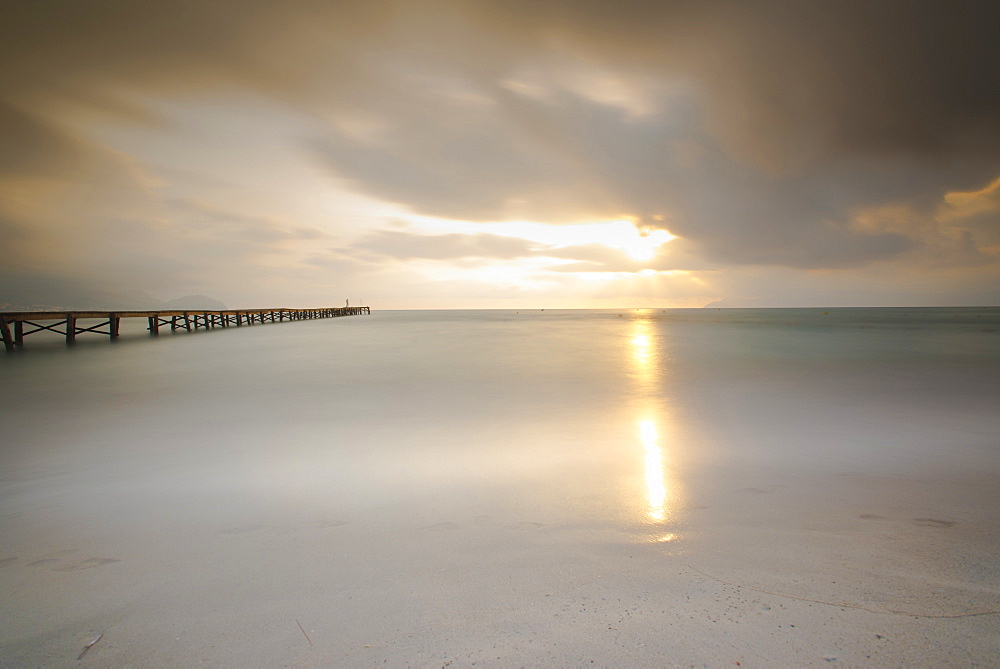 Long exposure of a wooden pier on Playa de Muro beach in the morning mood. The sky is overcast and fog lies over the Bay of Alcudia, Alcudia, Mallorca, Balearic Islands, Spain