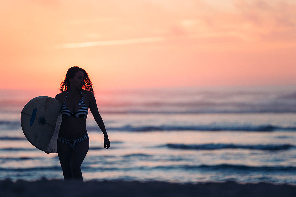 Female surfer goes with surfboard on the beach in sunset, surfing, Portugal, sunset