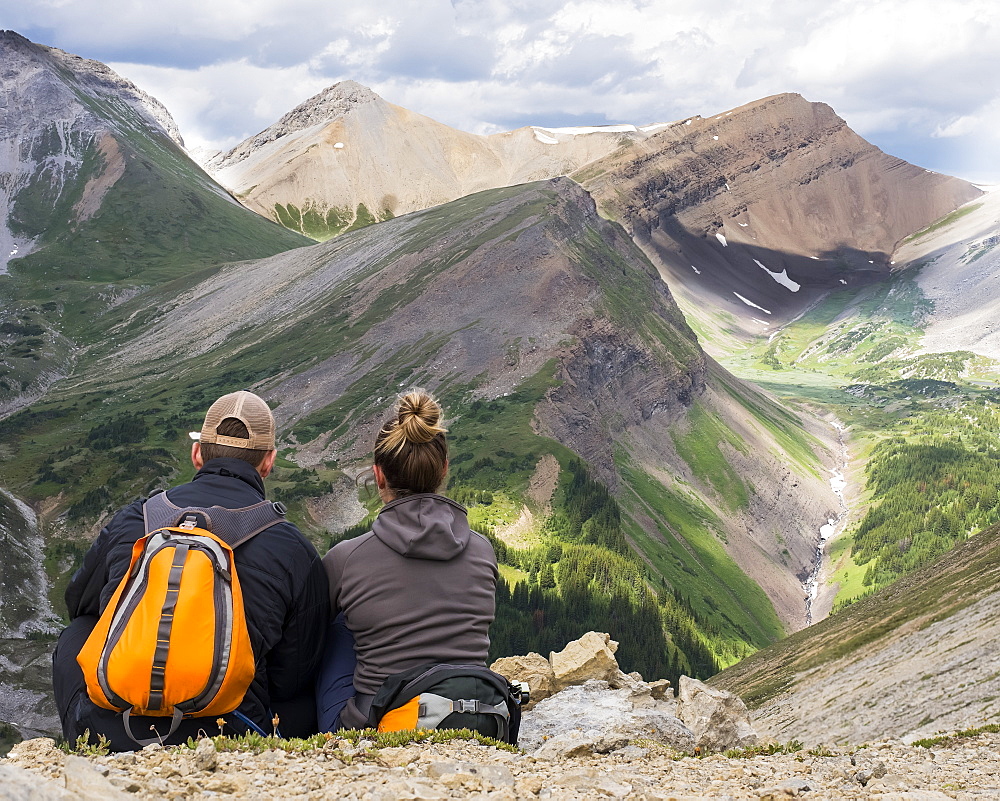 Male and female hiker sitting on a rock ridge overlooking a valley and mountain range in Kananaskis Country, Alberta, Canada