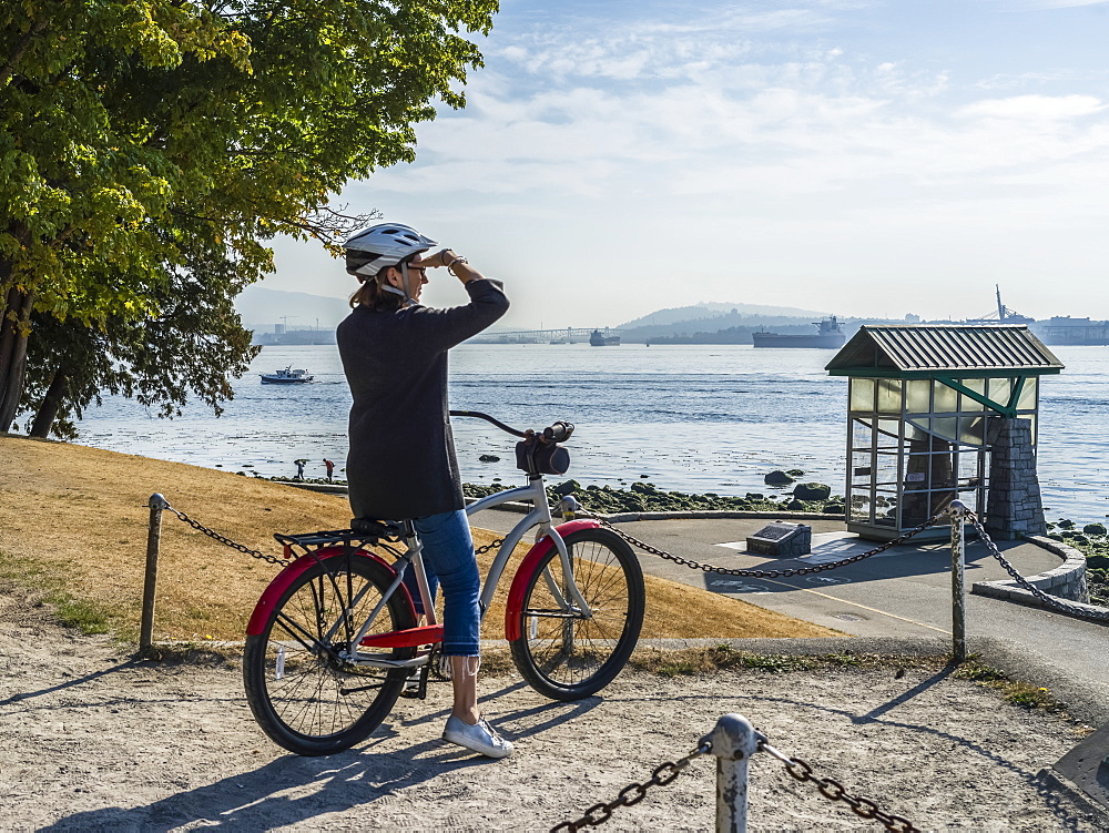 A woman sits on a bike looking out to the ocean and coastline at the Stanley Park Seawall, Vancouver, British Columbia, Canada